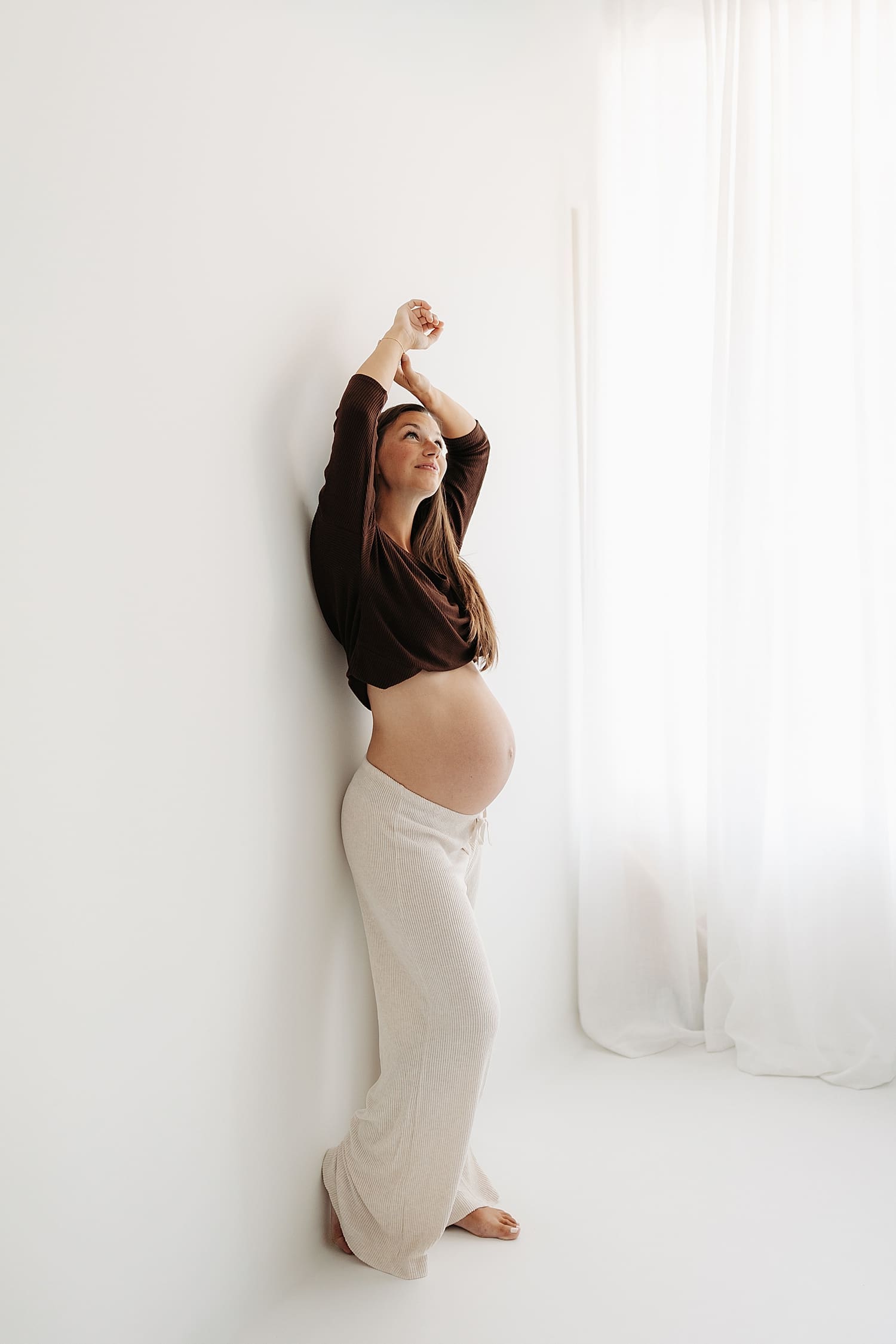 brunette pregnant mama stands against wall with hands up for studio maternity shoot