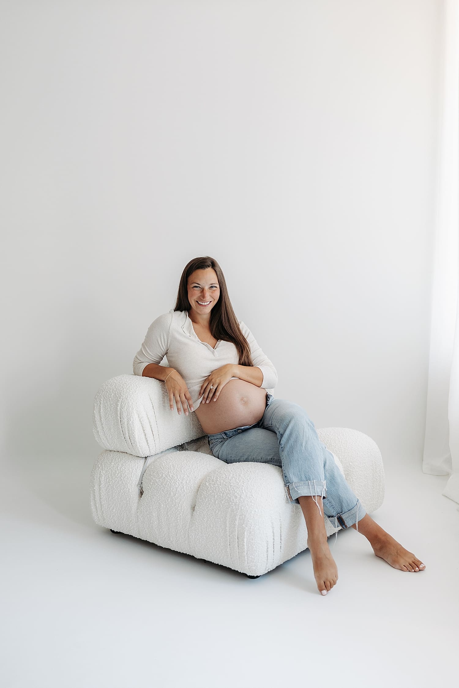brunette mom sits on puffer white chair for studio maternity shoot