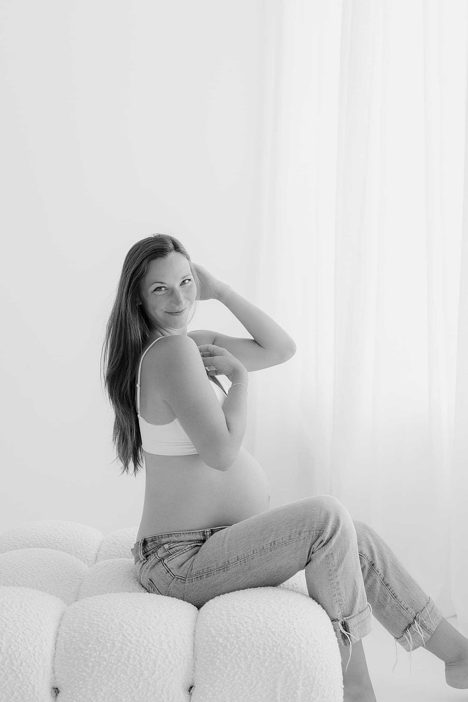 woman in bra and denim sits on puff coach for studio maternity shoot