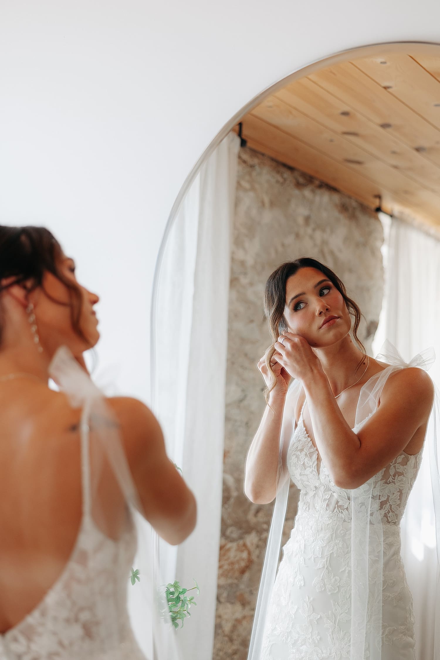 brunette bride adjusts her earrings in a mirror by Rose and Oak Photography