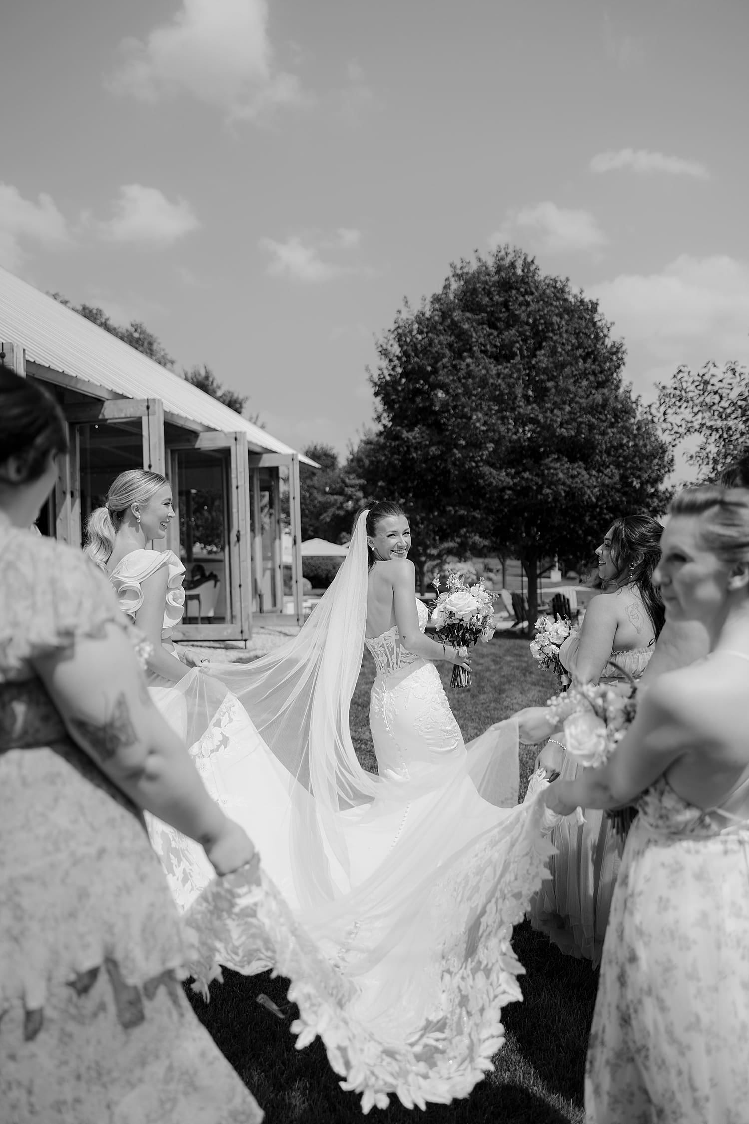 bride looks over her shoulder as bridesmaids lift her train by Rose and Oak Photography