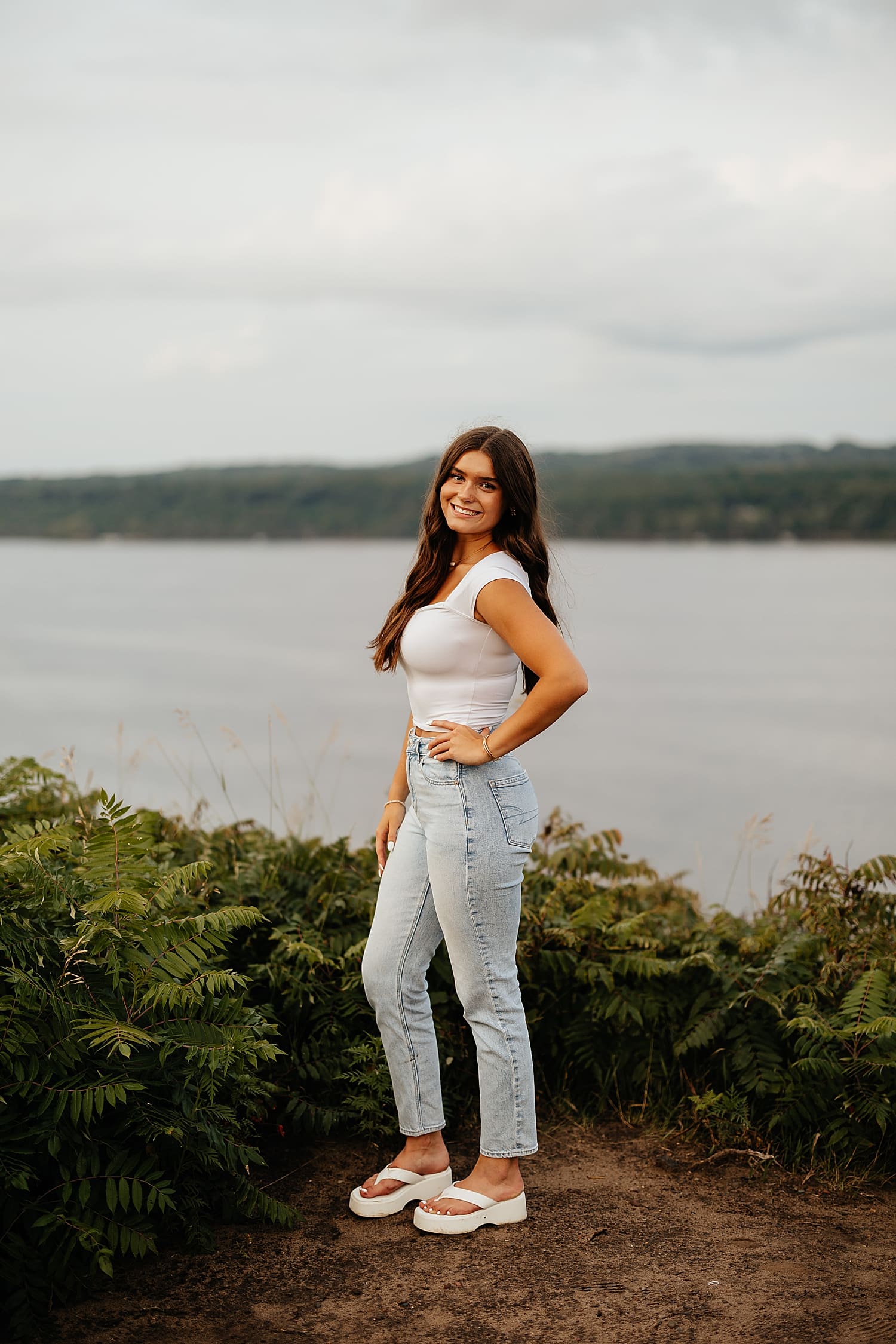woman in white shirt and denim stands in front of water for golden hour senior session