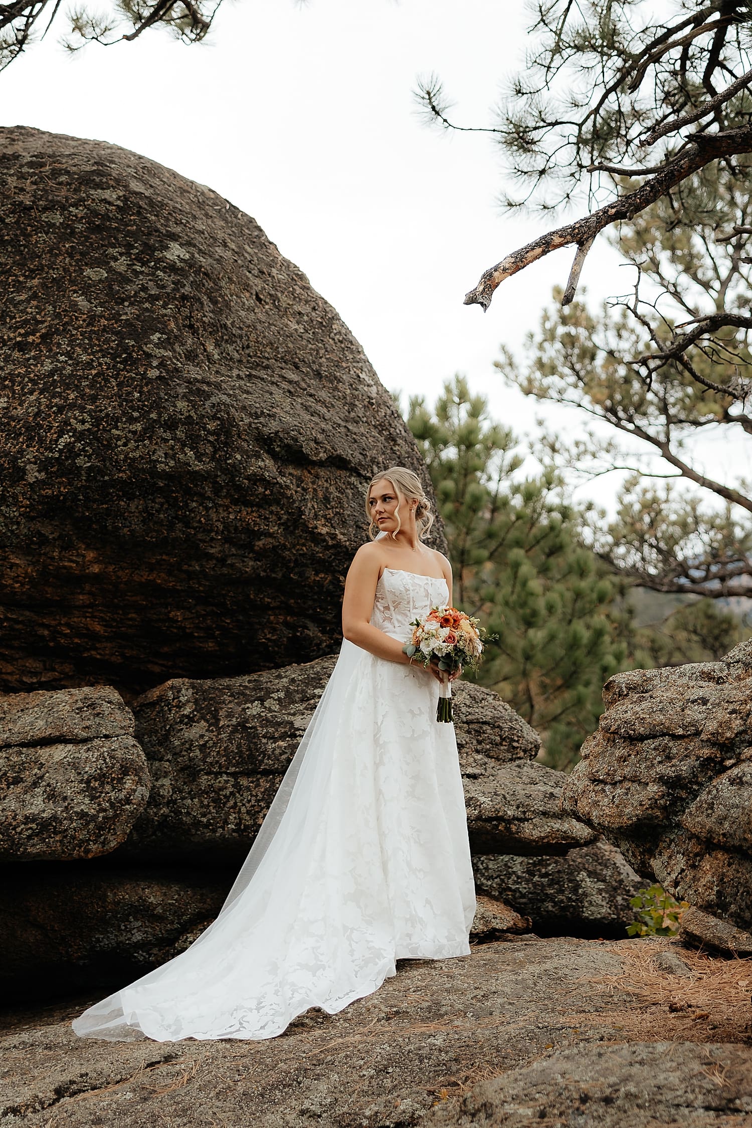 bride stands in white dress on large rock at laid-back celebration at Estes Park