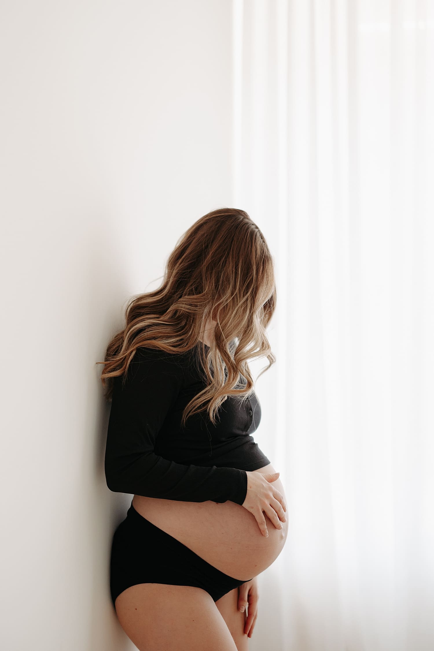 mother in black tee and underwear leans against white wall by Rose and Oak Photography