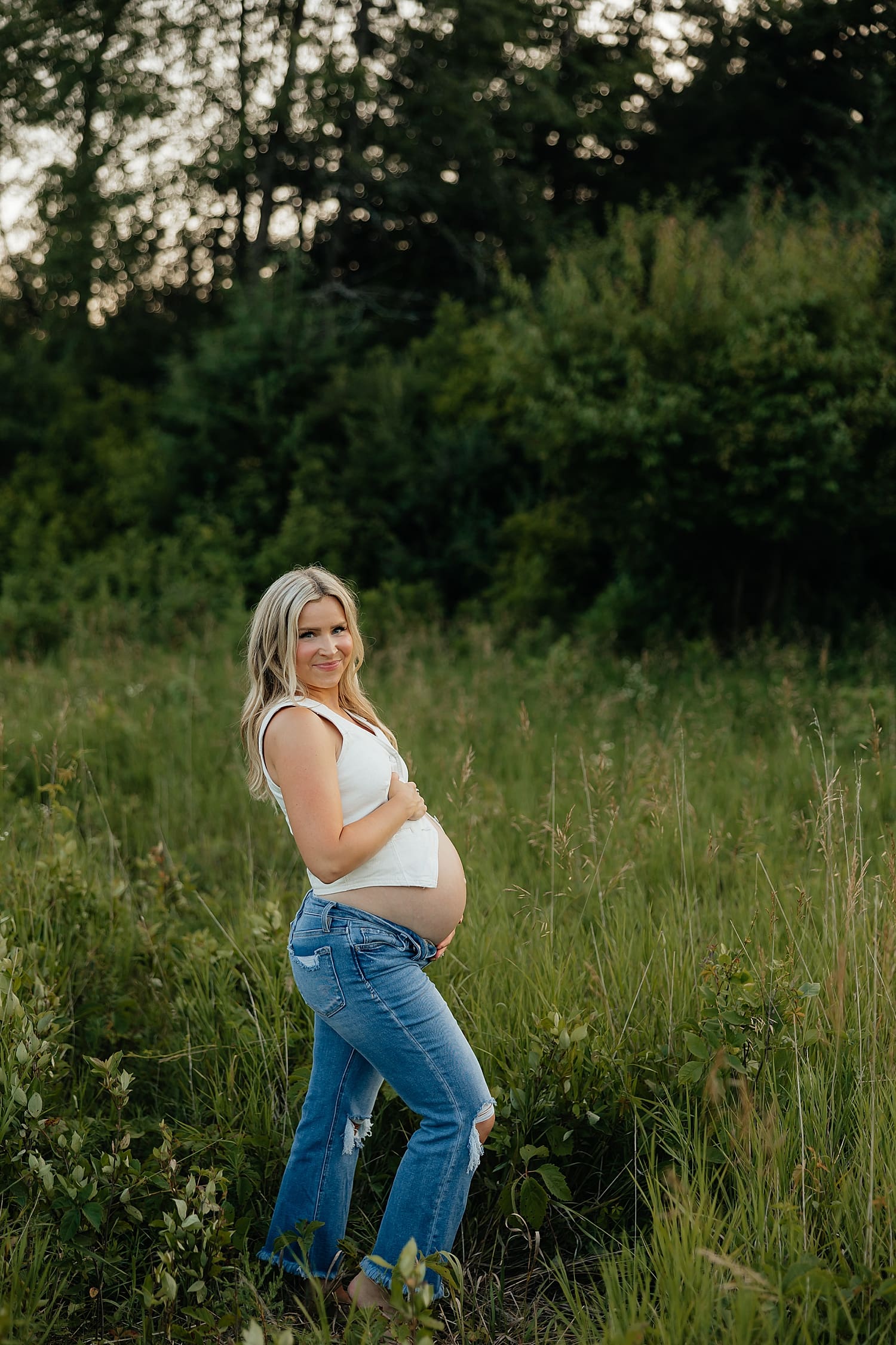 blonde expecting mother in jeans stands in tall grass by Rose and Oak Photography